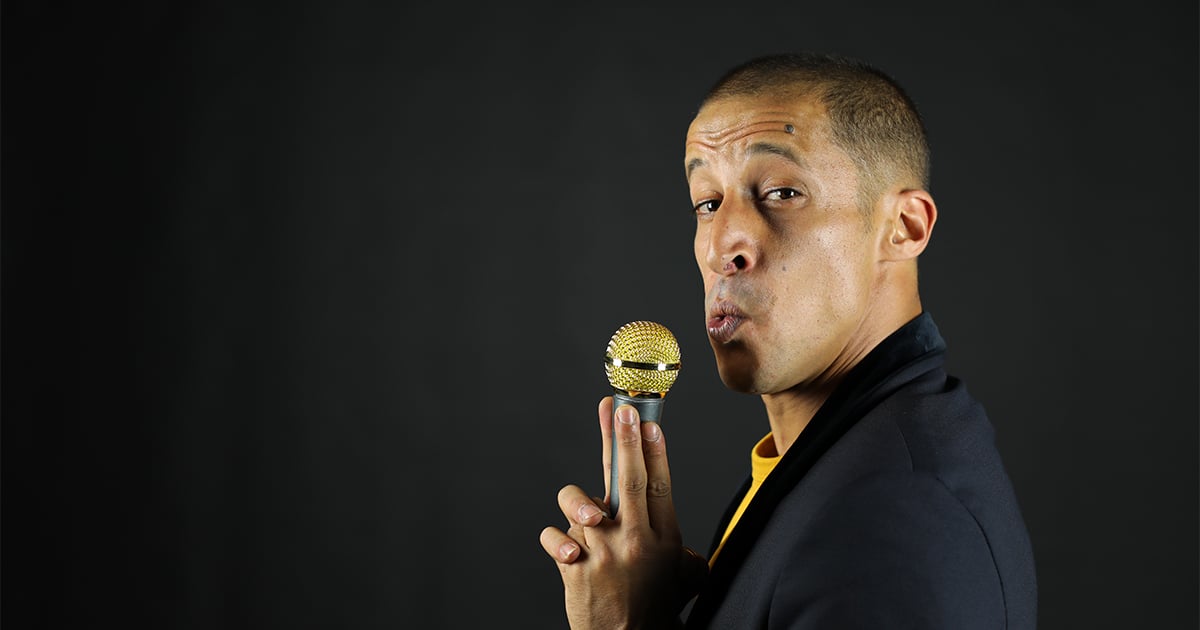 Kosh fixant l’objectif, bouche déformée, micro en main, doigts formant des pistolets.