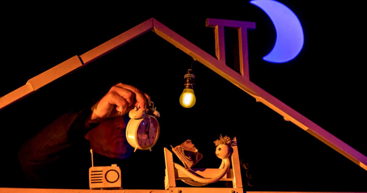 Un homme tenant un réveil face à une marionnette allongée dans un lit, dans une maison de poupée.